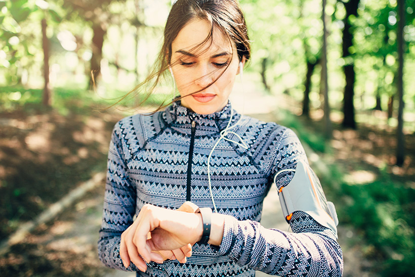 Frau mit Kopfhörer im Ohr joggt durch den Wald und schaut auf ihren Fitness-Tracker